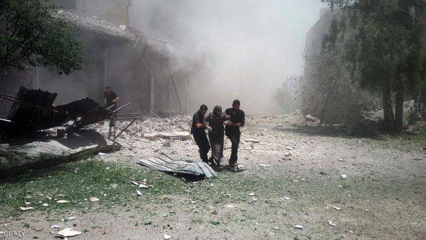 TOPSHOT - A cloud of dust is seen after a building collapsed as a Syrian woman is helped to leave the area following reported air strikes on June 11, 2016 in the town of Kfar Batna, on the outskirts of the capital Damascus. / AFP / AMER ALMOHIBANY        (Photo credit should read AMER ALMOHIBANY/AFP/Getty Images)