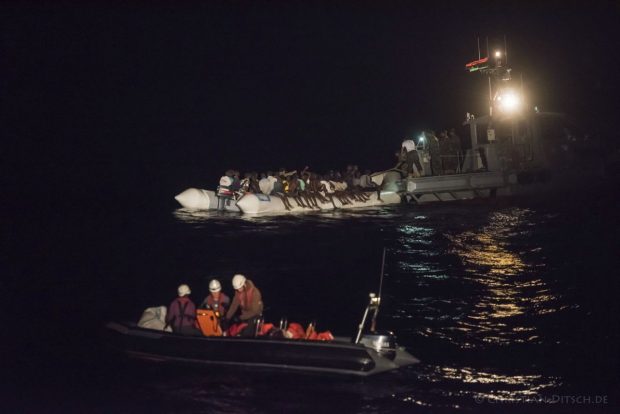 In this Oct. 21, 2016 picture provided by Sea-Watch aid group, refugees sit in a inflatable boat, background left, while a speedboat, labelled as Libyan Coast Guard, background right, and Sea-Watch members in a boat foreground arrive off the Libyan coast in the Mediterranean Sea. The German aid group Sea-Watch urged the European Union on Tuesday Oct. 25, 2016 to reconsider its plans to train Libyan forces to conduct sea rescue operations after a vessel labeled as belonging to the country's coast guard attacked the dinghy full of migrants last week. Dozens of people were feared dead in the incident. (Christian Ditsch/Sea-Watch via AP)